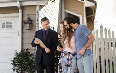 An agent in a suit showing a clipboard to a couple on the driveway of a weatherboard house