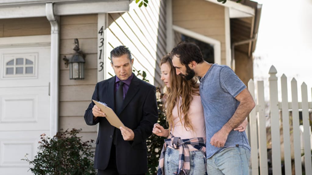 An agent in a suit showing a clipboard to a couple on the driveway of a weatherboard house